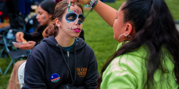 A woman paints colorful Day of the Dead face art on another woman outdoors.