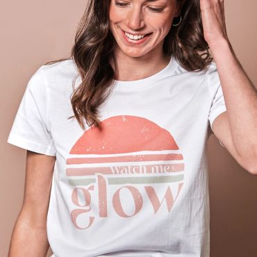 Woman wearing a white t-shirt with a retro sunset design and the words 'watch me glow'.