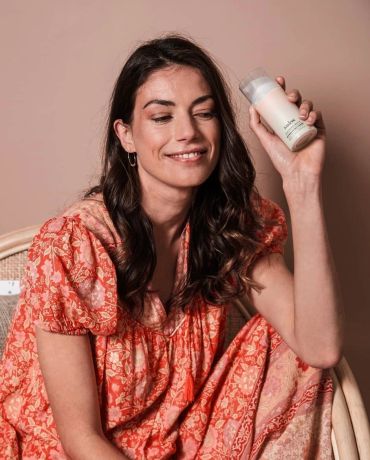 Woman in a floral dress holding a skincare product, smiling.