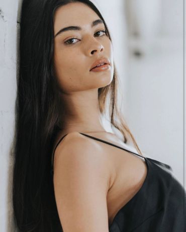 Woman with long dark hair leaning against a wall.