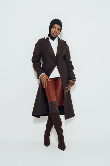 Fashionable woman in a long brown coat, leather pants, and boots, posing confidently.