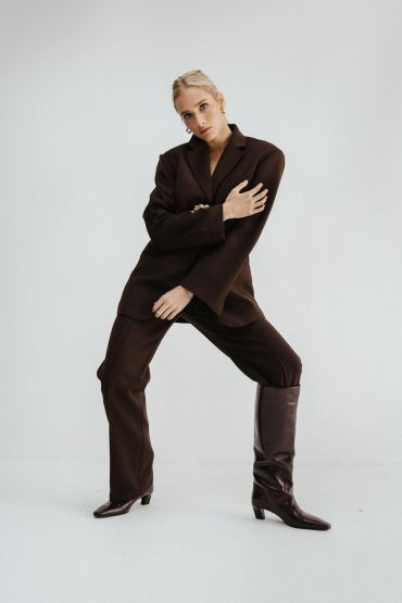 Woman posing confidently in a brown suit and boots against a plain background.