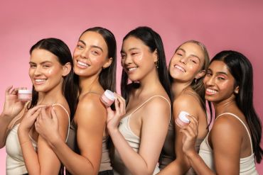 Five diverse women smiling and holding skincare cream jars against a pink background.