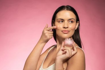 Woman applying moisturizer to her face against a pink background.