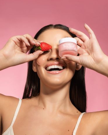 Woman holding a strawberry and skincare cream jar against a pink background.