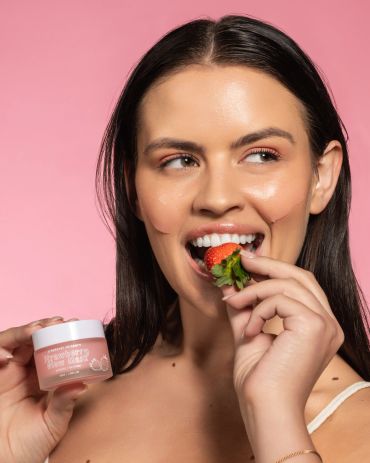 Woman enjoying a strawberry while holding a skincare product with facial patches on.