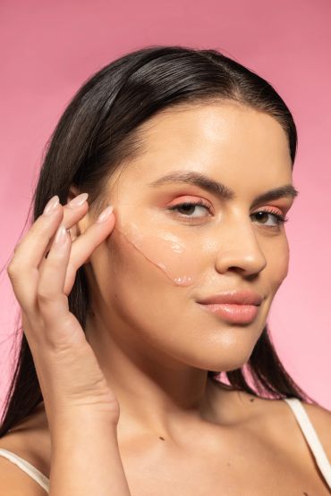 Woman applying skincare serum on her cheek against a pink background.