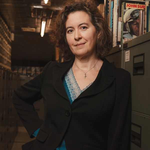 A confident woman stands in an archive room filled with files and magazines.