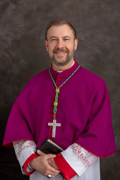 Clergyman in purple robe holding a book and wearing a cross necklace.