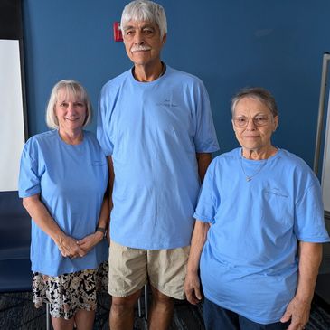 Three adults wearing light blue t-shirts stand indoors.
