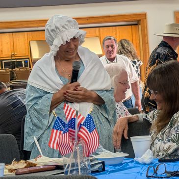 Woman in colonial attire speaking to seated guests at a patriotic event.