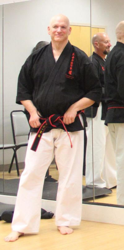 A man in a black martial arts uniform with a red and black belt, standing barefoot in a dojo.