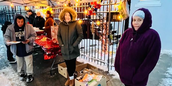 People bundled up at an outdoor winter food distribution under a tent with string lights.