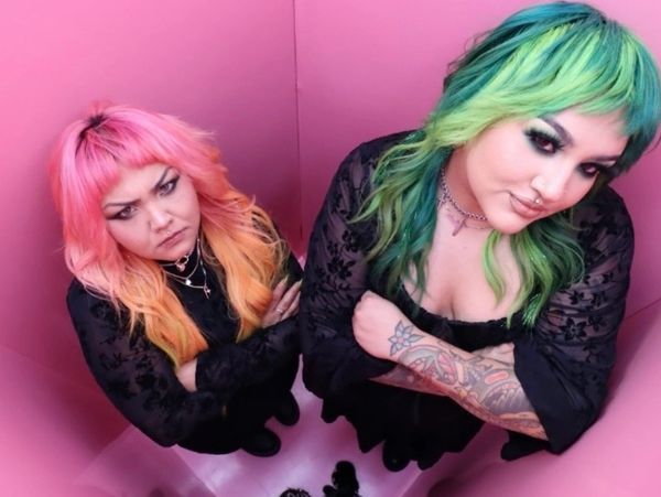 Two women with colorful hair and black outfits in a pink room, looking up.