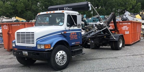 Blue and white roll-off truck with containers and scrap in background.