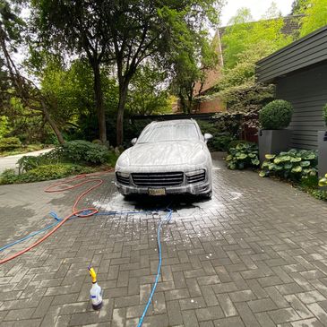 A car covered in soap foam during a wash in a driveway surrounded by greenery.