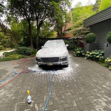 A car covered in soap foam during a wash in a driveway surrounded by greenery.