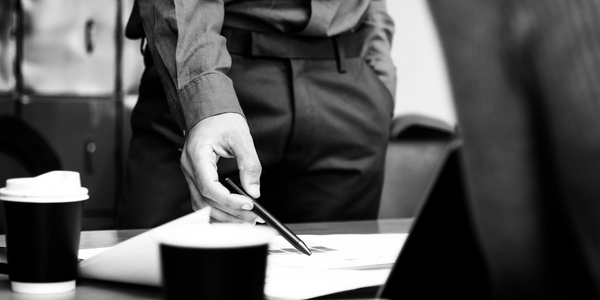 Black and white image of a person pointing at documents on a table with a pen.