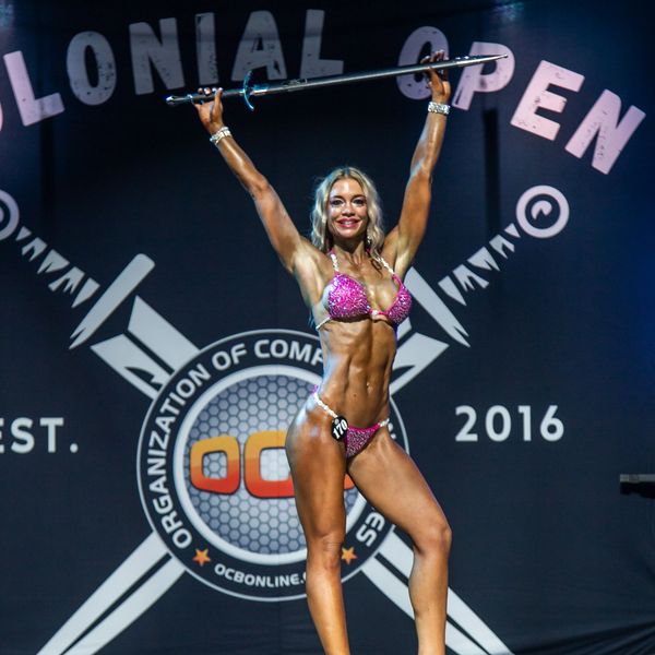 Female bodybuilder in a sparkling pink bikini poses on stage holding a sword overhead.