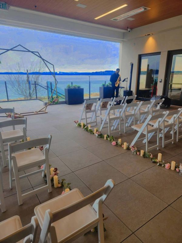 Indoor wedding setup with white chairs, flower decorations, and a saxophonist by the window.