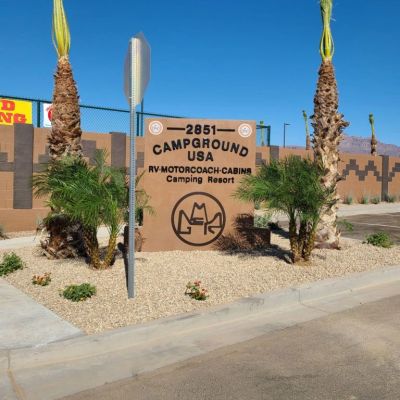 Entrance sign for Campground USA with palm trees and clear blue sky.
