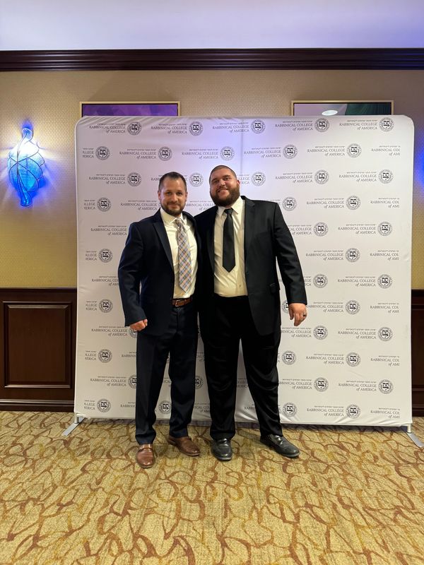 Two men in suits pose in front of a Rabbinical College of America backdrop.