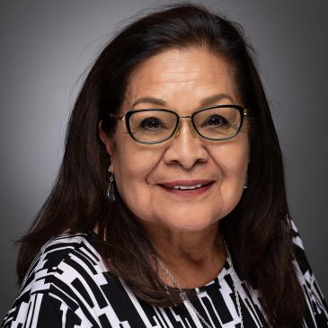 Smiling woman with glasses and patterned top against a gray background.