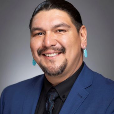 Smiling man in a blue suit with turquoise earrings.