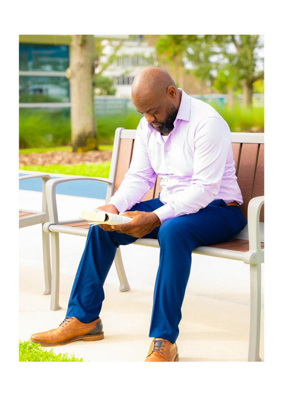 Man in dress shirt reading a book on a bench outdoors.