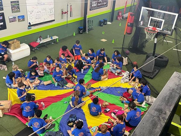 Group of children sitting in a circle on a colorful parachute indoors.