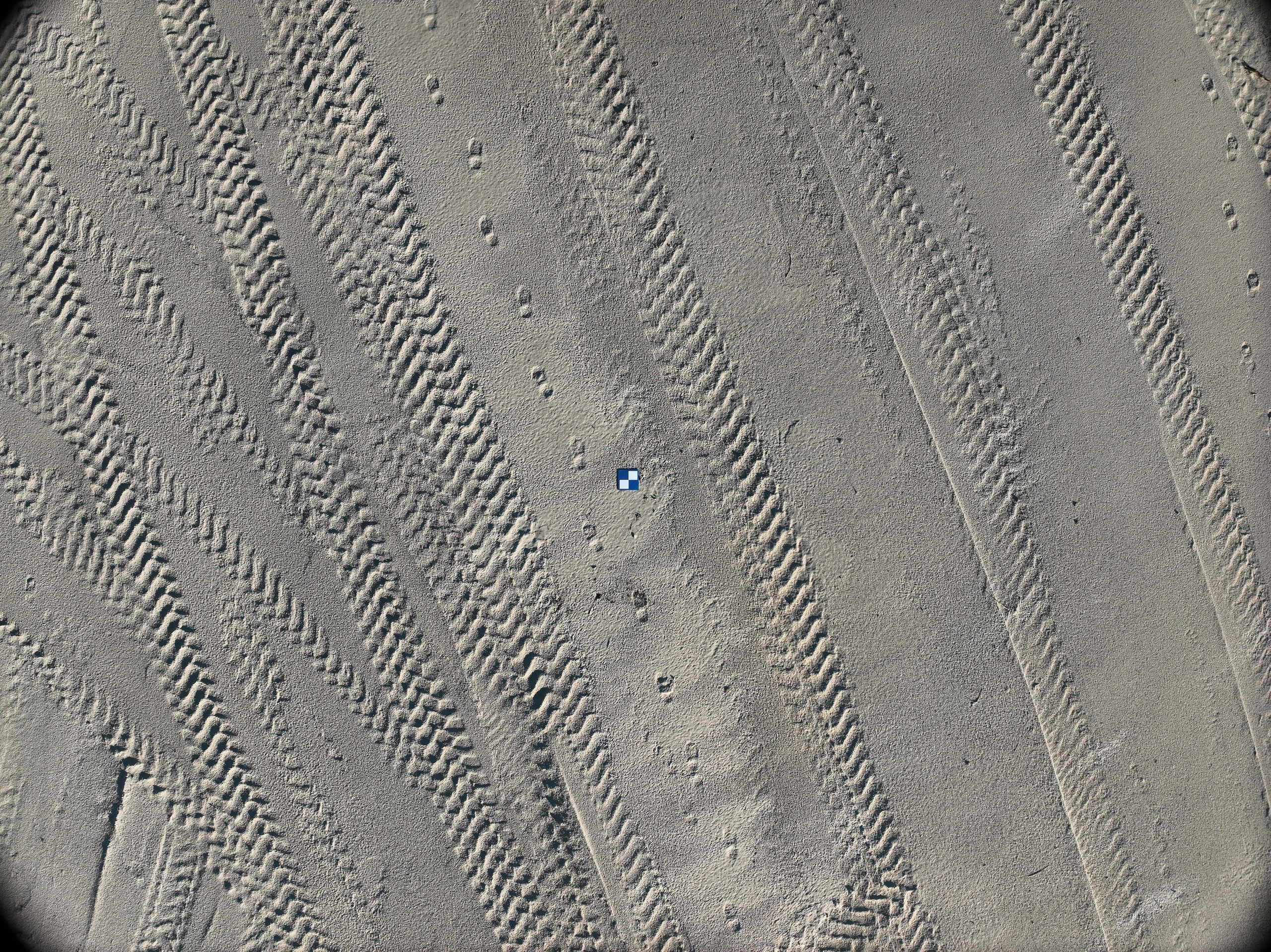 Tire tracks on a sandy surface with a small blue and white square object.