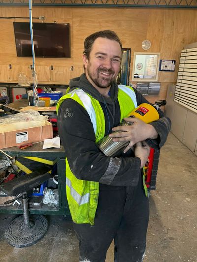Man in a high-visibility vest smiles holding a tool in a workshop.
