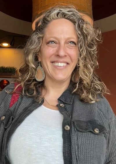 Smiling woman with curly hair wearing a gray jacket and large earrings.
