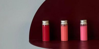 Five small bottles with different shades of pink and red on a dark red semi-circular shelf.