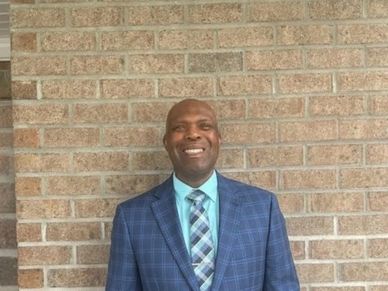 Smiling man in a blue plaid suit standing against a brick wall.