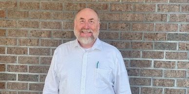 Smiling man in a white shirt standing against a brick wall.