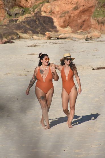 Two girls walking at Byron Bay beach, smiling and wearing the same one-piece terracota swimwear.