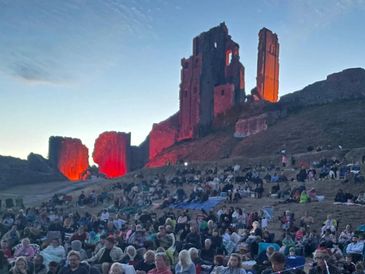 Crowd gathered at sunset near illuminated ancient ruins on a hill.