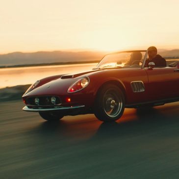 A classic red convertible sports car driving at sunset by the water.