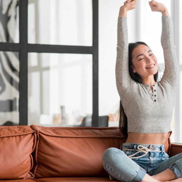 A young woman stretches happily on a brown leather couch in a bright room.