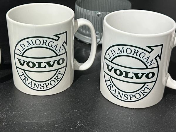 Two white mugs with J.D. Morgan Volvo Transport logo on a black surface.