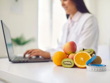 A woman works on a laptop with fresh fruits and a measuring tape nearby.