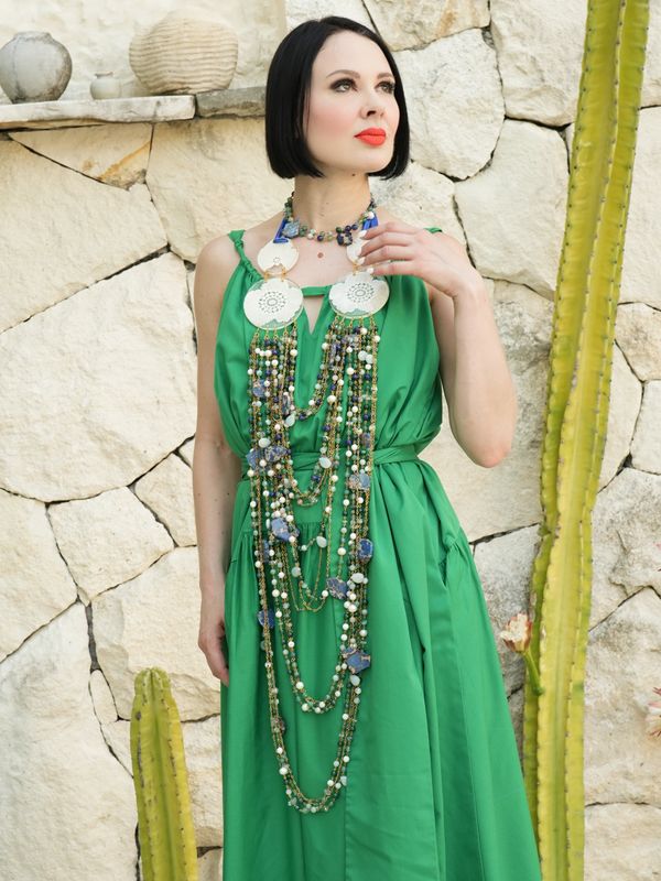 Woman in a green dress with elaborate jewelry poses by a stone wall and cactus.