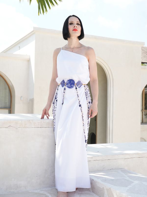 Woman in a white one-shoulder dress with blue embellishments posing barefoot outdoors.