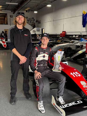 Two men in a racing garage, one holding a trophy.