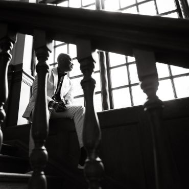 Man in a suit sitting by a large window, seen through stair railing in black and white.