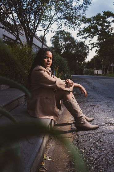 Woman in beige blazer and boots sitting on a roadside curb.