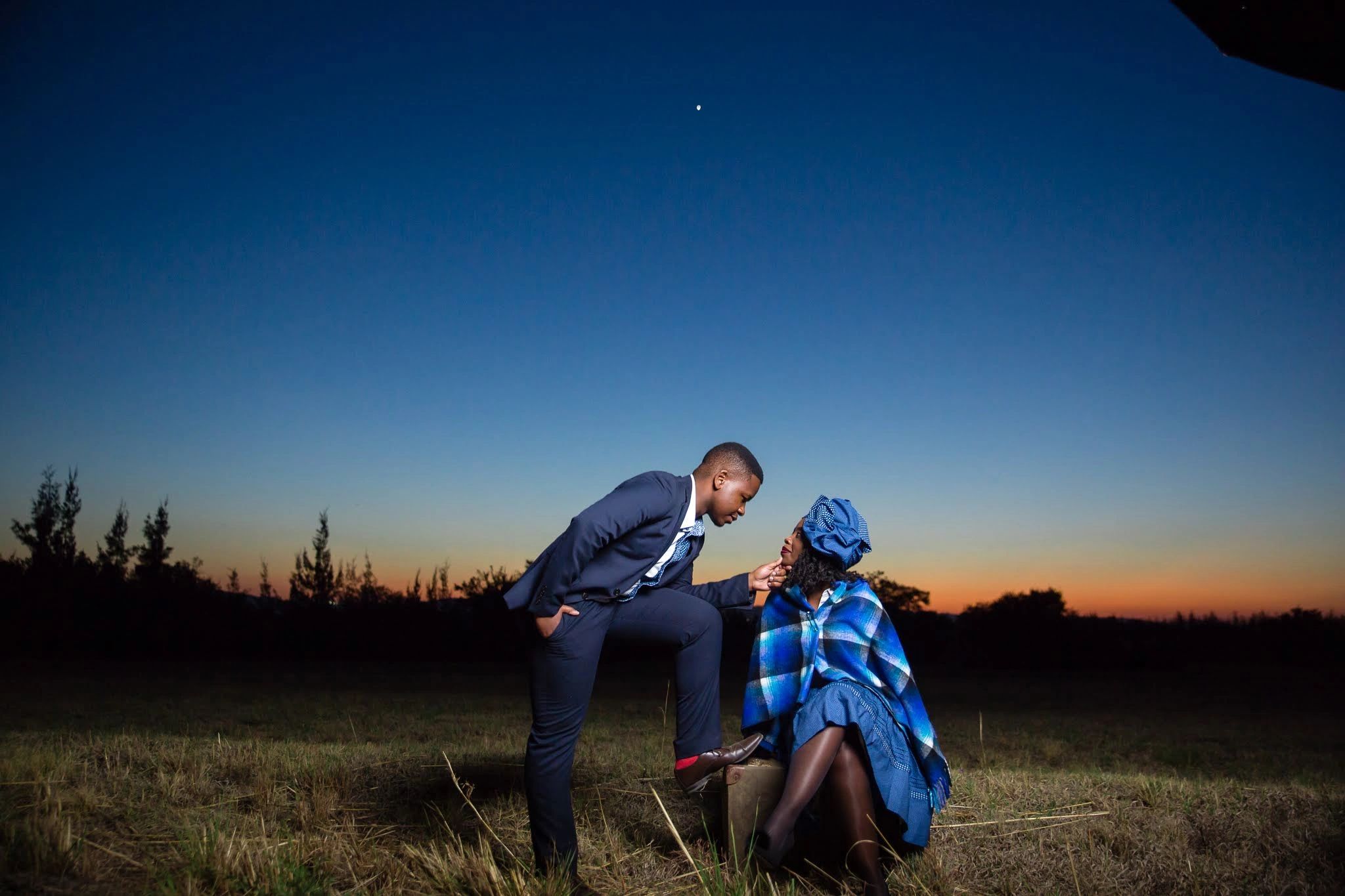 A couple shares a tender moment outdoors at sunset with a clear sky.