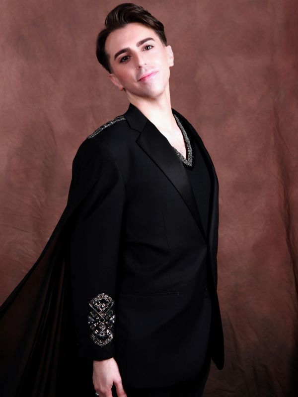 A man posing in a stylish black suit with embellishments.