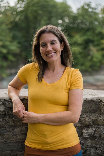 white woman with brown hair wearing a yellow shirt stands by a stone fence with trees in the backgro